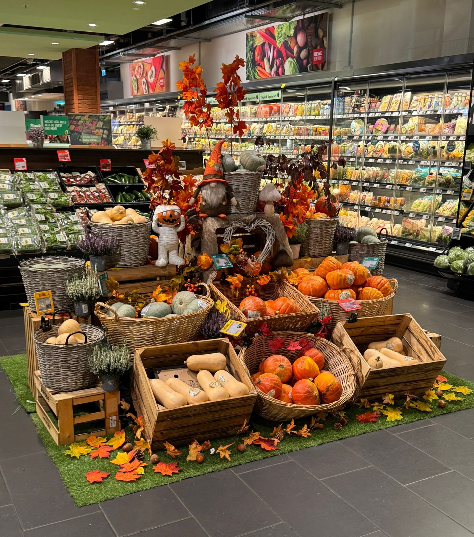 Herbstlich dekorierte Präsentation mit verschiedenen Kürbissorten, Holzkisten, Körben, bunten Blättern und Figuren im Obst- und Gemüsebereich.